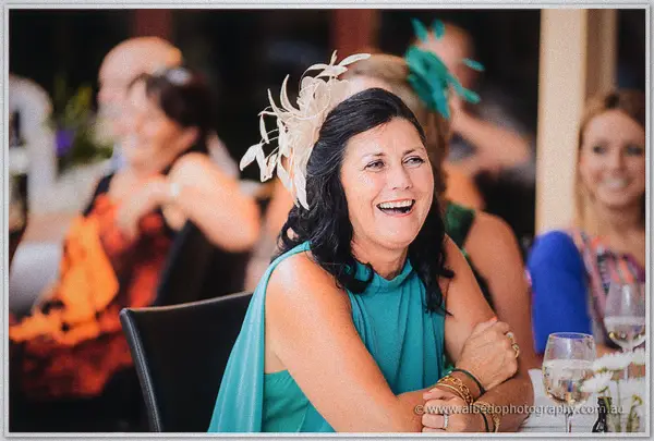 A guest in a teal dress and cream fascinator laughs mid-speech among smiling tablemates