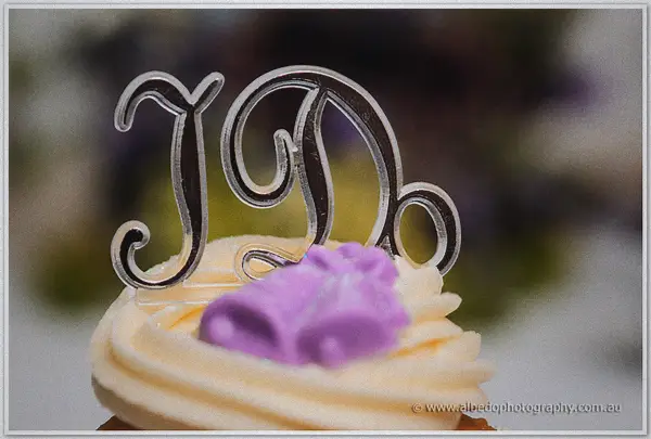 An I Do topper with purple bells sits on a swirl of buttercream atop a vanilla cupcake