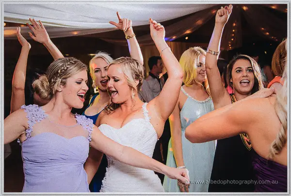 The bride in a white gown dances with a bridesmaid in lavender, arms raised, on a packed dance floor