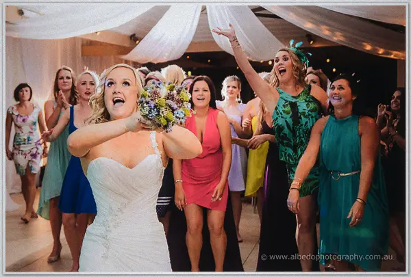 Bouquet toss moment: the bride turns back mid-throw as eager guests surge forward under the marquee
