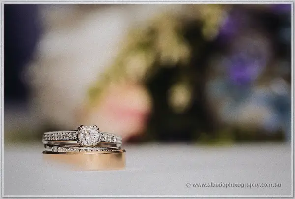 A diamond solitaire stacked with wedding bands gleams on white, the bouquet a soft blur behind
