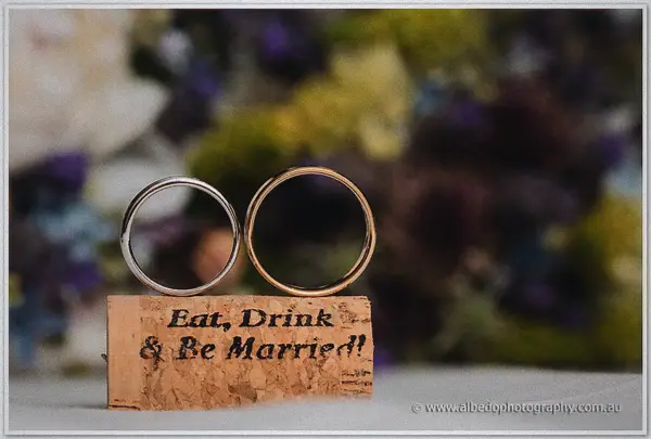 Two wedding bands stand upright on a cork stamped Eat, Drink and Be Married, blooms blurred behind