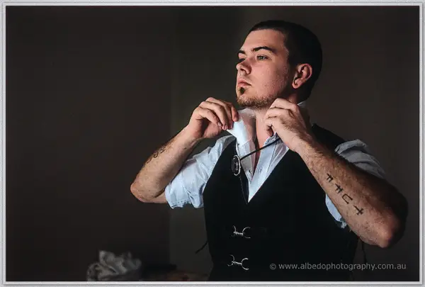 Gary adjusts his collar in moody sidelight, tattooed forearms and dark vest, in the farm kitchen