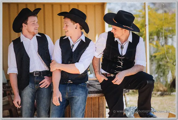Three groomsmen in black hats and vests laugh together beside a timber bench outside the shearing quarters
