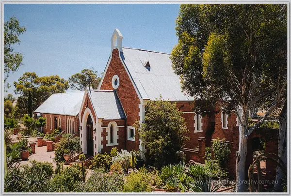 Red-brick bush chapel with white trim and tin roof rises among gum trees and garden beds