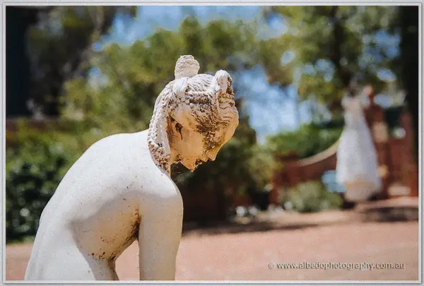 Weathered garden statue bows before the red brick chapel, Tash's white gown blurred on the path beyond