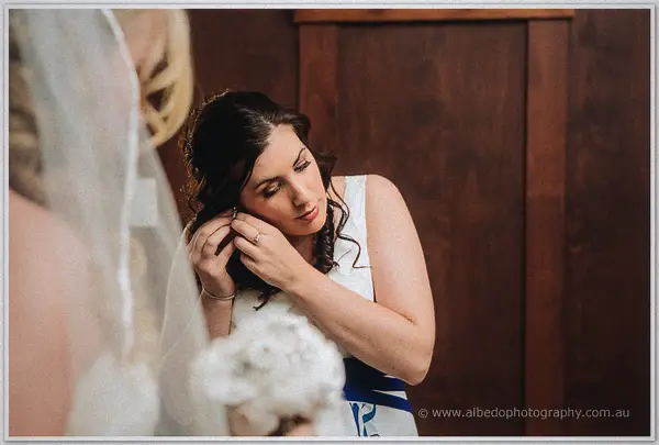 Bridesmaid in white fastens an earring while Tash waits veiled in the timber-lined room