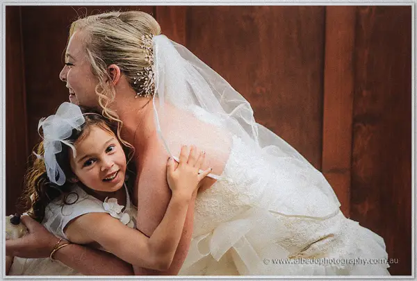 A flower girl in a white dress hugs the smiling bride beneath her veil against dark timber panelling.