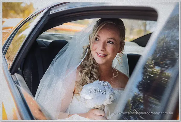 The beaming bride peeks from the back seat of the bridal car holding her white bouquet in soft shade.