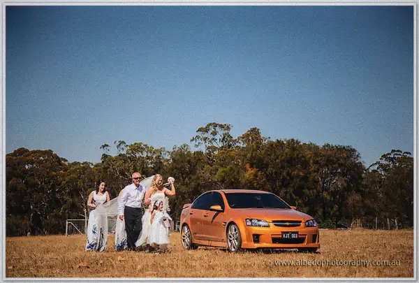 Tash and Gary walk their bridal party across a dry paddock toward an orange sedan under clear blue sky.