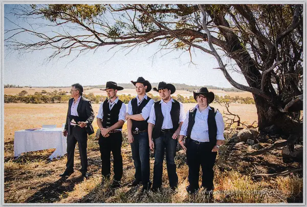 Groomsmen in black hats and vests line up under a gnarled tree as the celebrant waits beside a covered table.