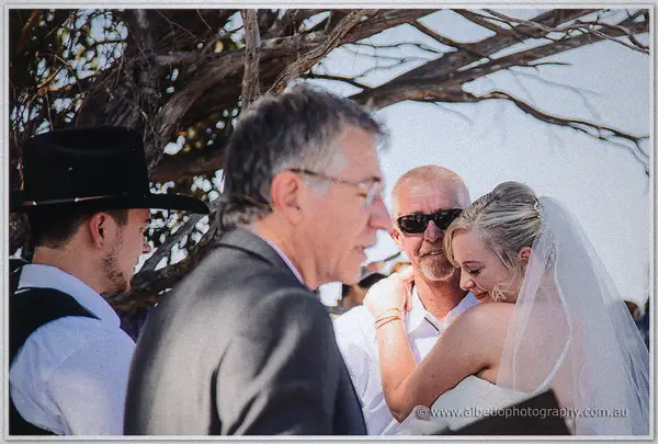 The bride embraces her father beneath the ceremony tree while guests and the celebrant look on.