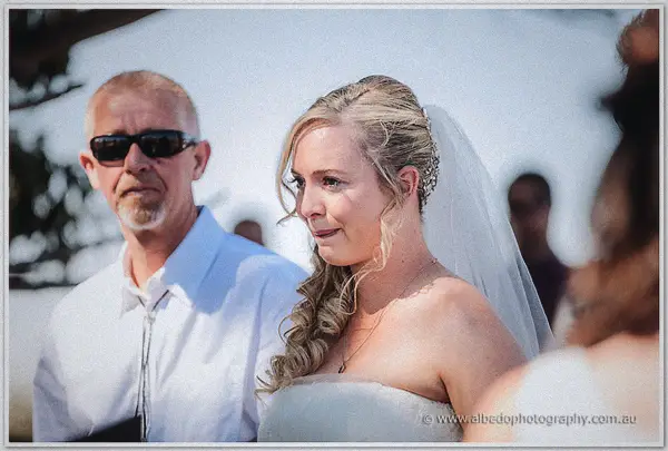 The bride watches the ceremony with a soft smile while an older man in sunglasses stands close by.