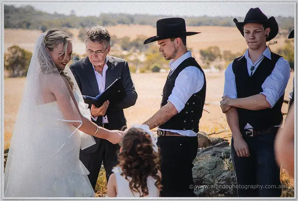 The celebrant reads from his folder as the bride and her hatted groom hold hands with a flower girl watching.