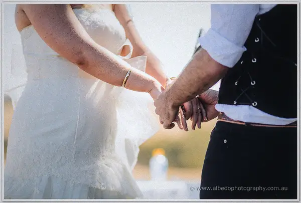 A close-up of the couple's joined hands and wedding rings against sunlit white lace and a dark vest.