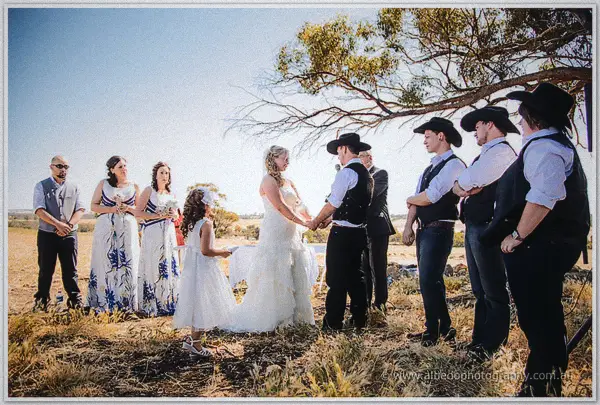 Guests in blue-print gowns and black hats circle the couple as they marry under wide country skies.