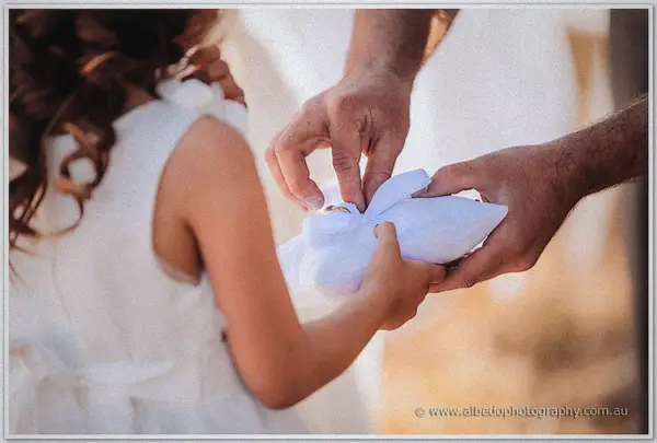 A flower girl's small hands steady the white ring pillow as the groom lifts a band from its bow.
