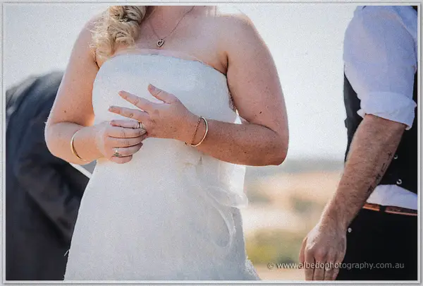 A close crop of the bride adjusting her new ring against sunlit lace, her groom's arm at her side.