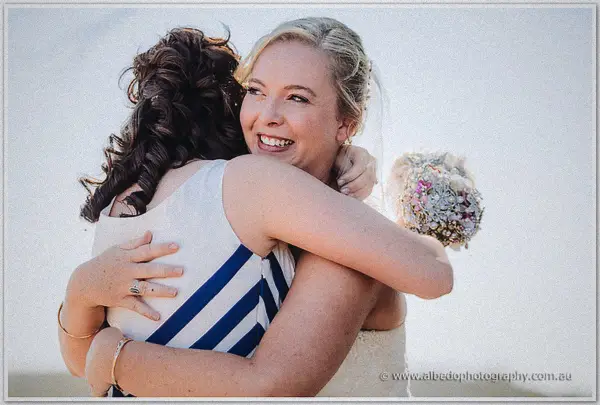 The bride hugs a dark-haired friend in a striped dress, her bouquet slung over one shoulder in bright sun.