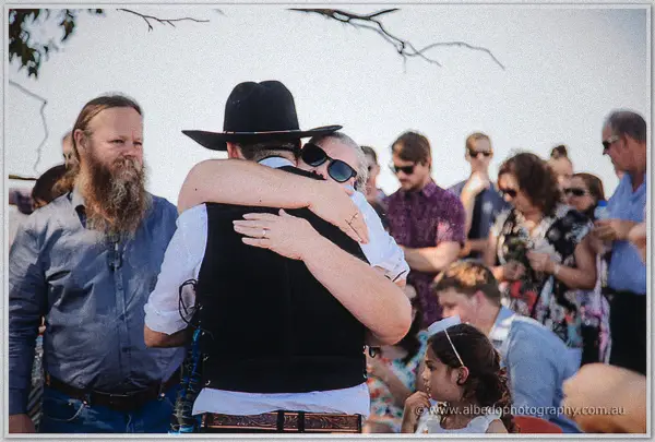 Gary hugs a guest in sunglasses amid a sunlit crowd of family and a small girl watching on.