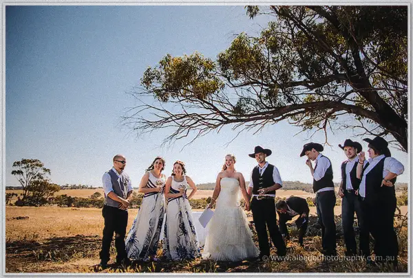 The full bridal party laughs together under a sprawling gum tree against brilliant blue outback sky.