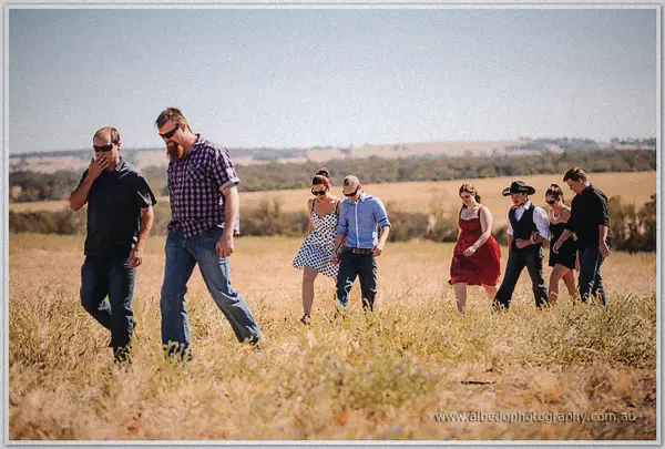 Guests in jeans, red dresses and hats trudge single file through waist-high golden grass under the sun.