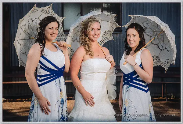 The bride and two bridesmaids in white-and-blue dresses pose with lace parasols before the weathered shed.