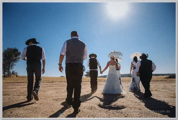 The bridal party strides away along a dusty track, parasols and hats silhouetted against the blazing sun.