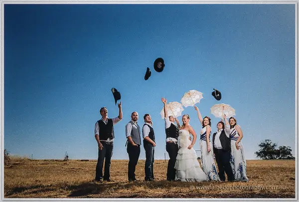 Groomsmen fling their black hats skyward as the bride and parasol-twirling bridesmaids cheer in the paddock.