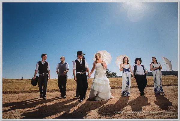 Tash and Gary lead their laughing party down the red dirt road, parasols raised against the afternoon sun.
