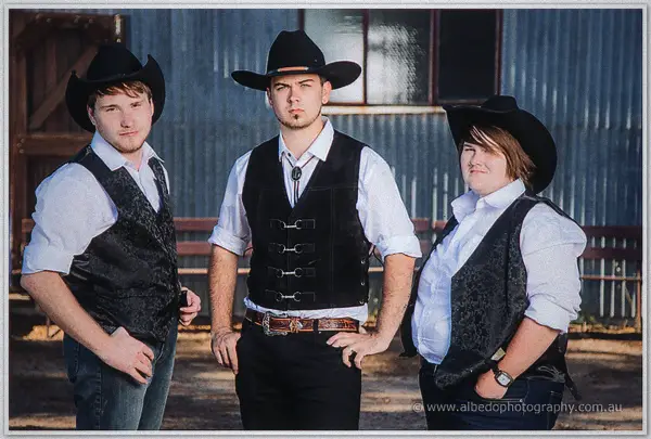 Three groomsmen in black vests and cowboy hats square up to the camera before the corrugated shed.