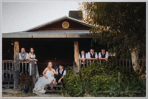 Tash and Gary sit on the homestead steps flanked by their wedding party at the veranda railing