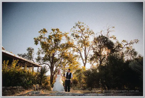 Tash and Gary walk hand in hand across a rocky rise, sunset flaring through the trees behind