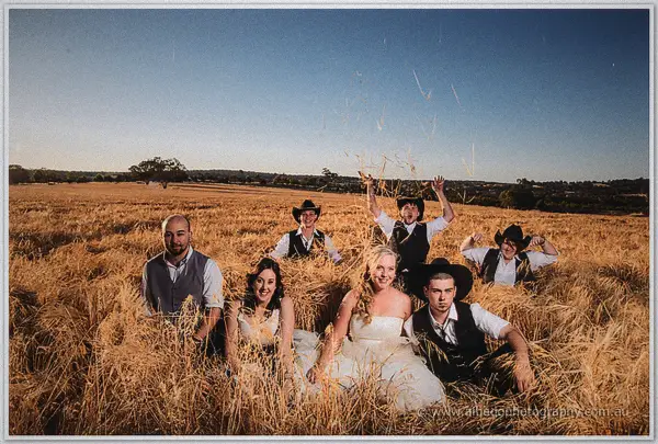 The wedding party lounges in the oat crop, groomsmen tossing straw skyward behind Tash