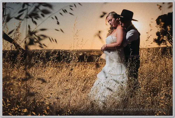 Gary wraps his arms around Tash from behind in a dusk field, seed heads framing the glow