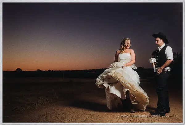 Tash gathers her flowing gown on a dirt track as Gary watches, holding her bouquet