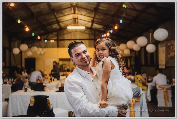 A groomsman in white cradles a laughing flower girl inside the festooned reception shed