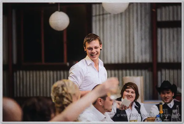 A laughing groomsman raises a toast between seated guests beneath the paper lanterns