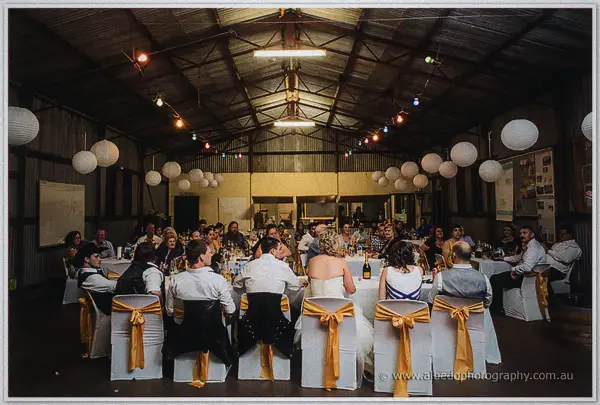Reception guests fill the lantern-lit shearing shed, gold sashes trailing down the trestles