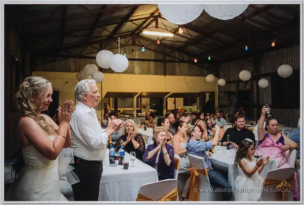 Tash and her father applaud beneath white paper lanterns as guests clap along in the shed