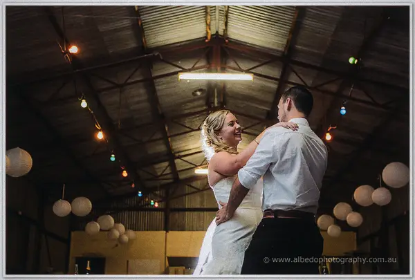 Tash and Gary dance beaming beneath coloured festoon bulbs and paper lanterns in the barn