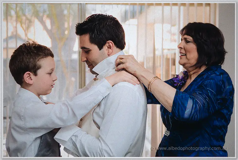 A page boy and Craig's mother adjust his collar as he dresses in soft window light