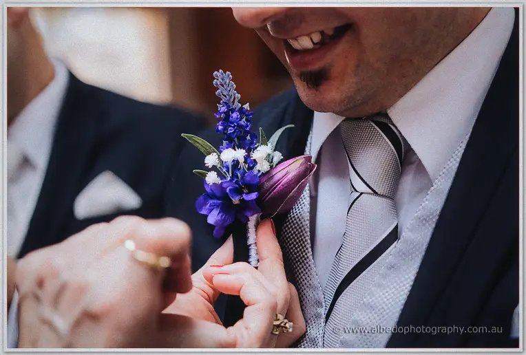 Smiling groom as hands fix his purple and plum boutonniere beside a patterned grey tie
