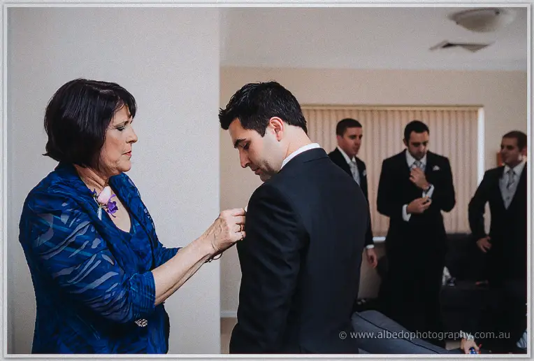 Craig's mother pins a buttonhole to his black suit while groomsmen dress in the background