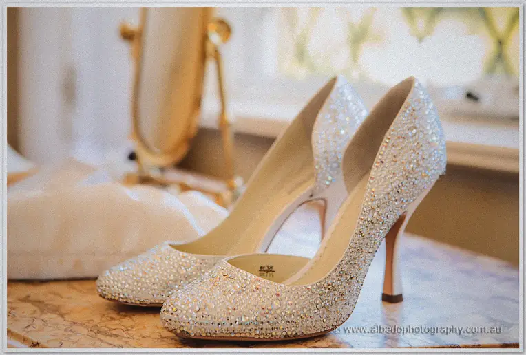Crystal-encrusted ivory bridal heels glow on a marble dresser by a sunlit window