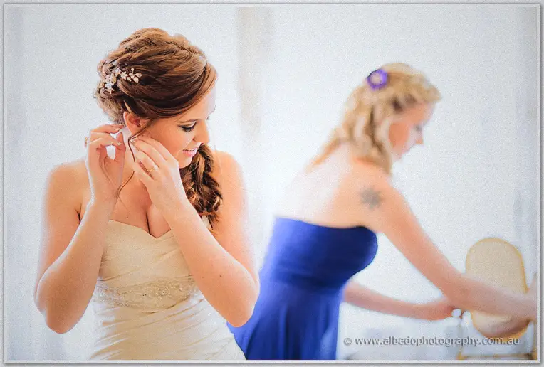 Amanda fastens an earring beside her updo as a bridesmaid works behind her