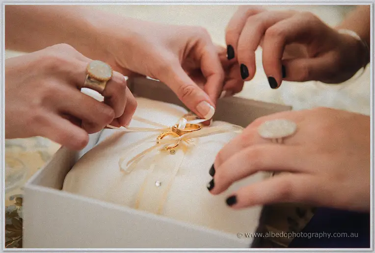 Bridesmaids' hands tying gold wedding rings to a satin ring pillow in a white box