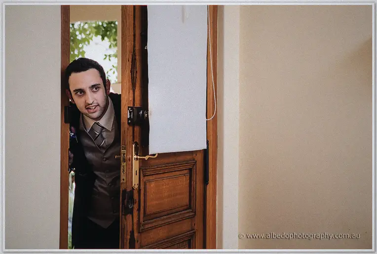 A grinning groomsman peeks around a half-open timber door of the bridal suite