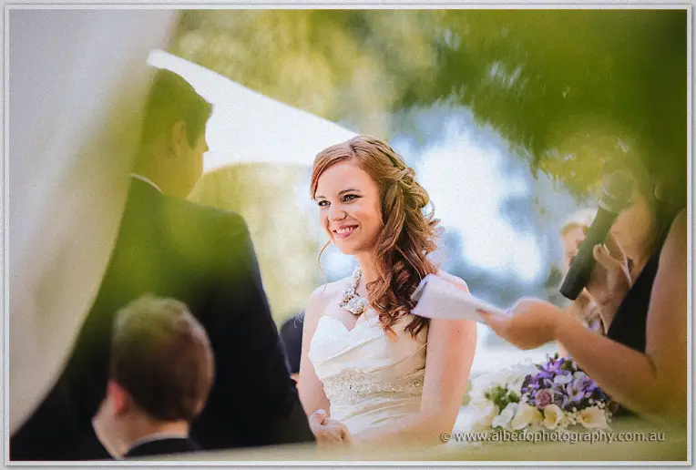 Amanda smiling at Craig as the celebrant reads beneath arbour drapery