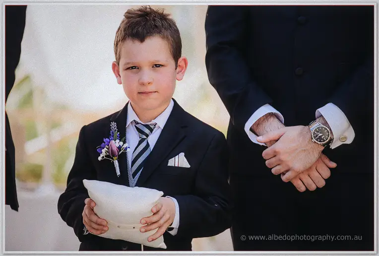 The page boy cradling the ring pillow beside a groomsman's crossed hands